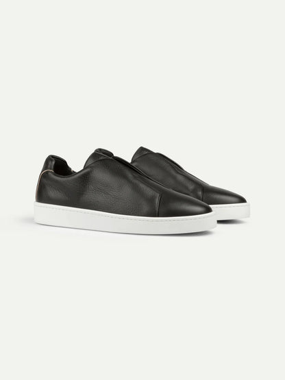 A pair of Aurélien Black Grain Leather Slip-On Sneakers with a minimalist design, smooth black leather, no visible laces, and thick white soles are positioned side by side on a plain white background. , available at Richard Gelding Mayfair