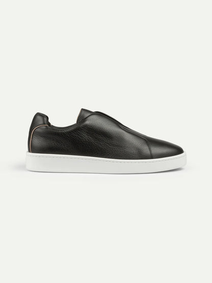 The Black Grain Leather Slip-On Sneaker by Aurélien features luxury pebbled leather, a minimalist design, rounded toe, elastic side panels, and a crisp white rubber sole. , available at Richard Gelding Mayfair