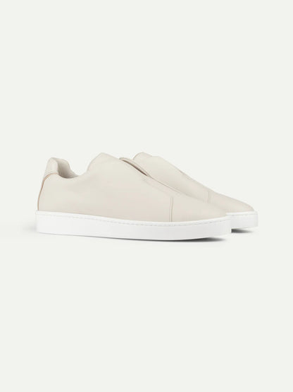 The Aurélien Ivory Grain Leather Slip-On Sneaker features a sleek, minimalist design with smooth off-white leather uppers and clean white soles. This luxury laceless shoe is shown on a plain light background with no visible laces or branding. , available at Richard Gelding Mayfair