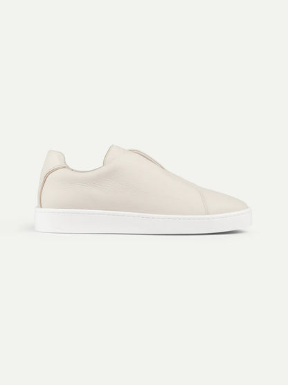 The Aurélien Ivory Grain Leather Slip-On Sneaker is a minimalist beige luxury laceless shoe with a smooth grain leather upper and clean white rubber sole, shown in side profile against a plain light background. , available at Richard Gelding Mayfair
