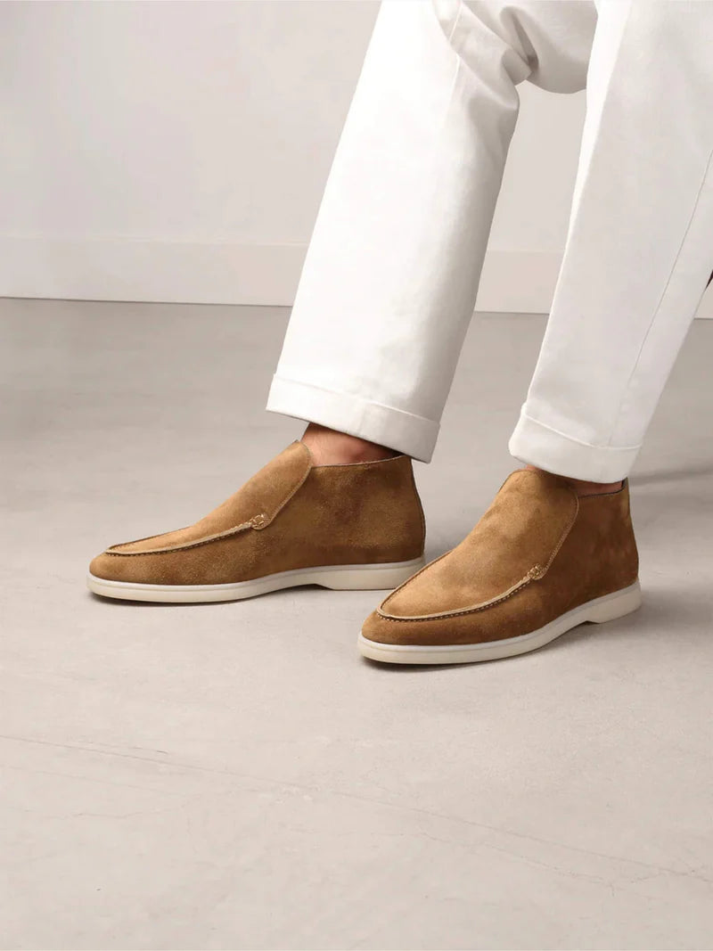 Indoors, a person in white pants showcases Aurélien's Beige Softey Suede City Loafer—smart luxury shoes with minimalist design and visible stitching—against a neutral grey floor and plain light wall background. , available at Richard Gelding Mayfair