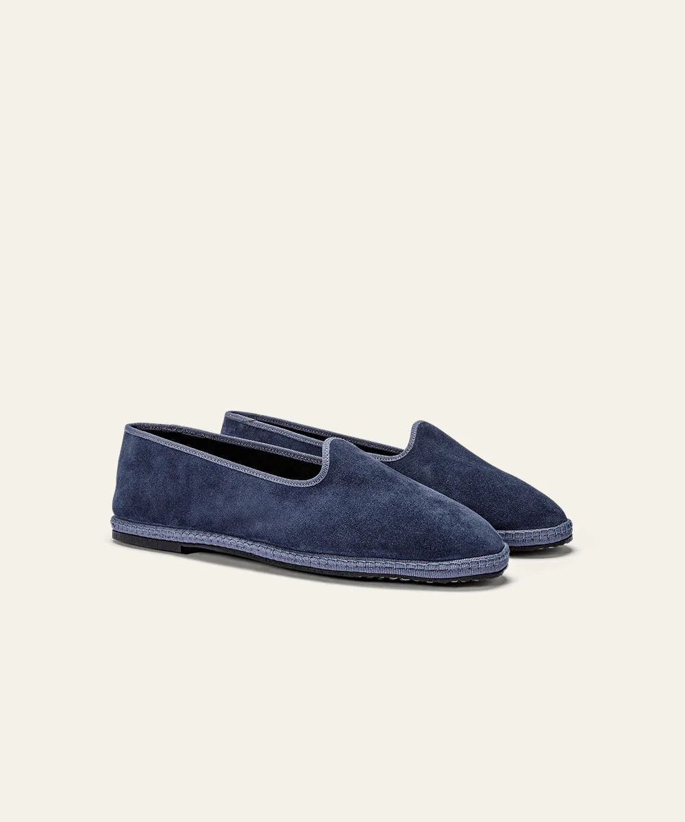 The Azaleri Space Blue Suede Friulane Certosa shoes feature soft suede in a seamless, elegant slip-on design with rounded toes and textured dark blue soles, shown angled on a neutral beige background for a refined casual look. , available at Richard Gelding Mayfair