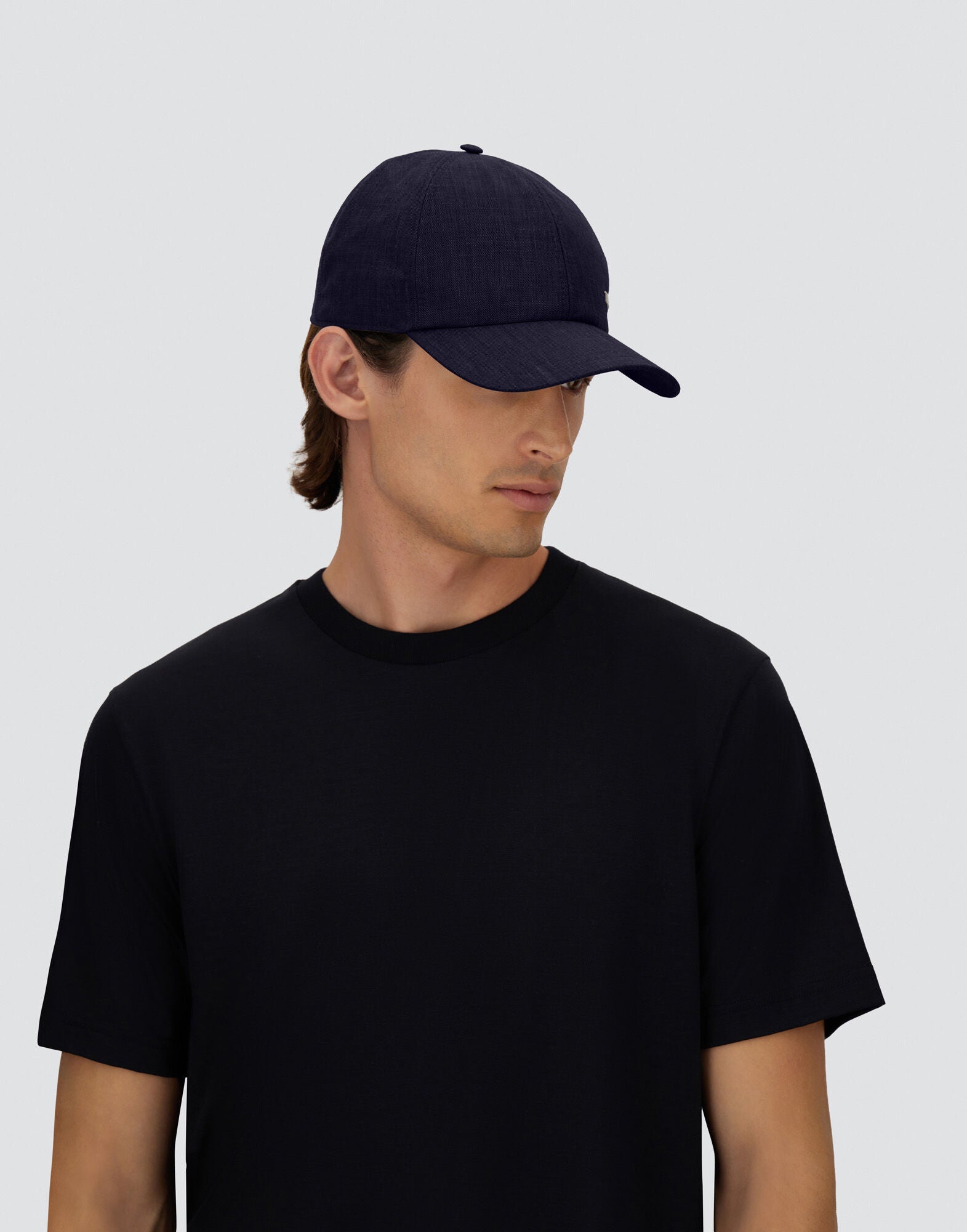 A person wearing a Herno Navy Wool/Silk/Linen Loro Piana Woven Cap and a plain black t-shirt stands against a light gray background, gazing slightly down and to the side, with medium brown hair tucked behind their ears and visible under the cap. , available at Richard Gelding Mayfair