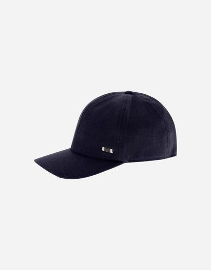 The Herno Navy Wool/Silk/Linen Loro Piana Woven Cap features a dark navy hue, curved brim, rectangular metallic logo on the left panel, visible stitching, and a smooth, structured shape on a light grey background. , available at Richard Gelding Mayfair