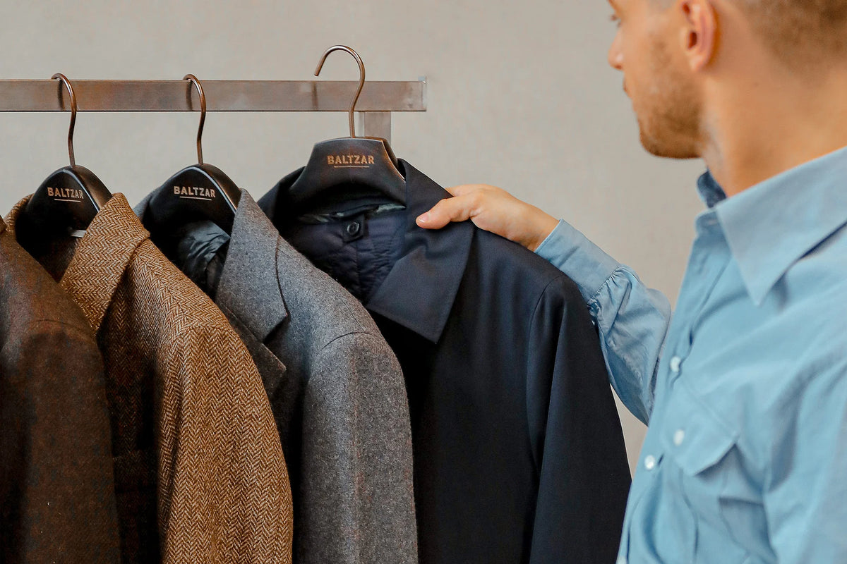 A man in a light blue shirt examines a row of four coats in various shades—brown, gray, and dark blue—hanging on a metal rack. Each coat is on a hanger labeled BALTZAR. The background is a plain, neutral wall. , available at Richard Gelding Mayfair