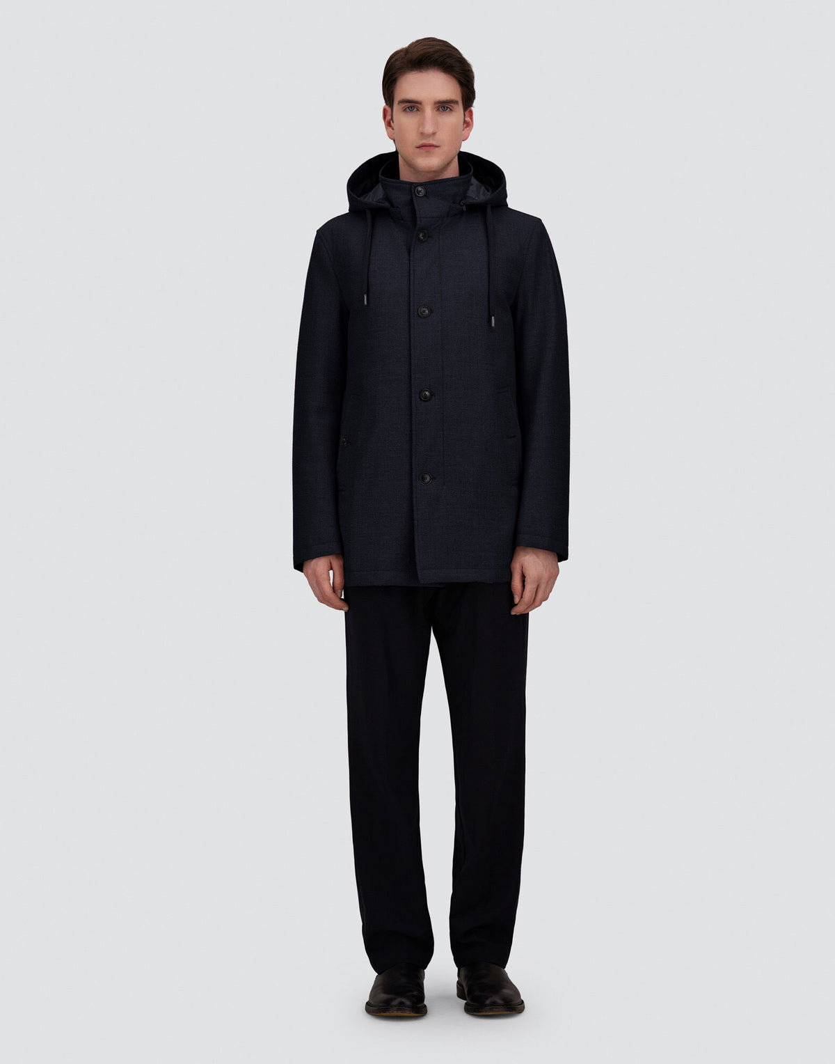 A young man stands facing forward against a plain gray background, wearing a navy blue Herno parka crafted from luxurious Loro Piana wool and silk, featuring a removable hood. He pairs it with dark trousers and black shoes. His arms rest by his sides and his expression is neutral. , available at Richard Gelding Mayfair