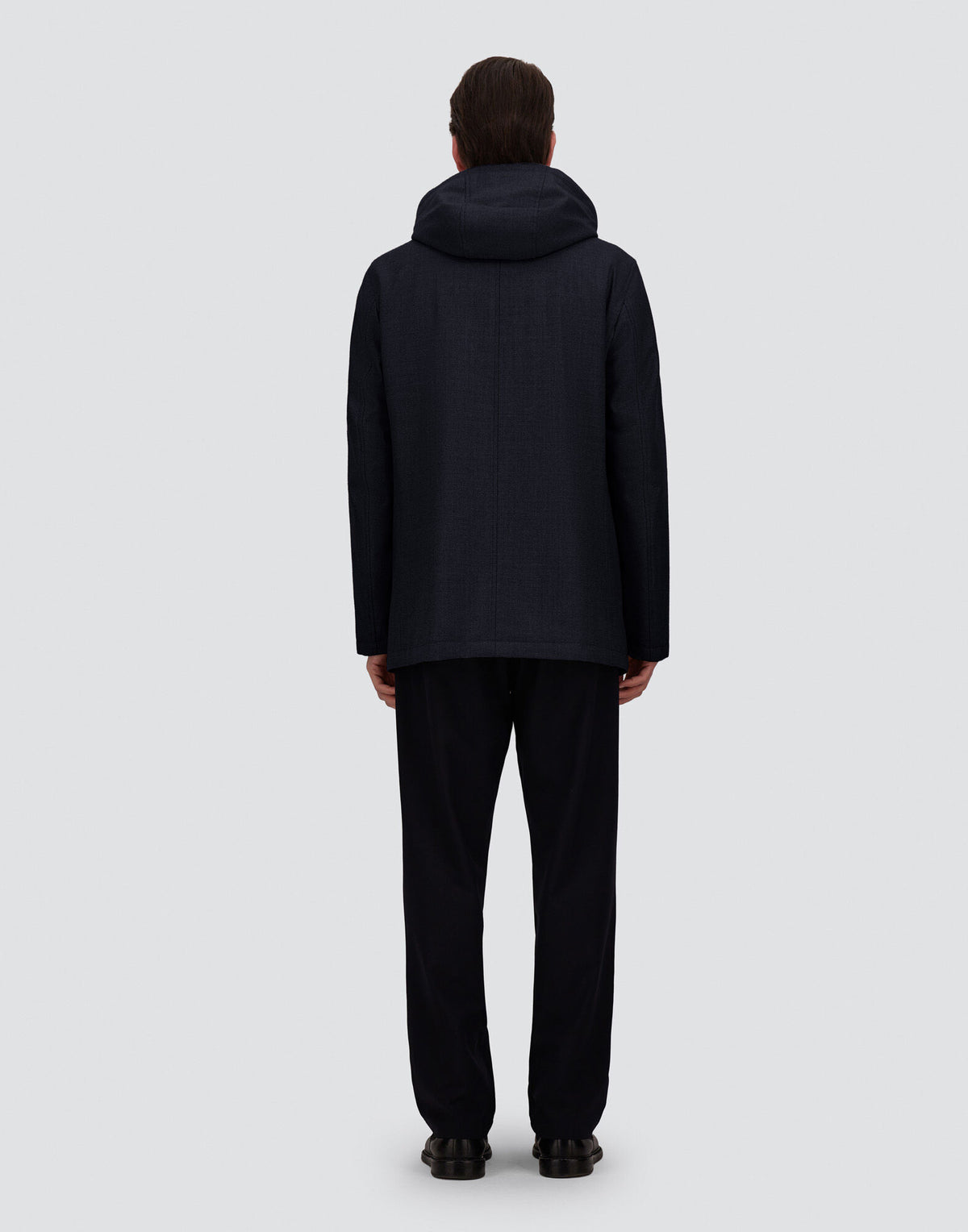 A person stands facing away from the camera, wearing a Herno Navy Blue Loro Piana Wool/Silk Parka with removable hood and matching dark pants. Their hair is short and neat, the background is plain light gray, both arms are relaxed at their sides, and they are wearing black shoes. , available at Richard Gelding Mayfair