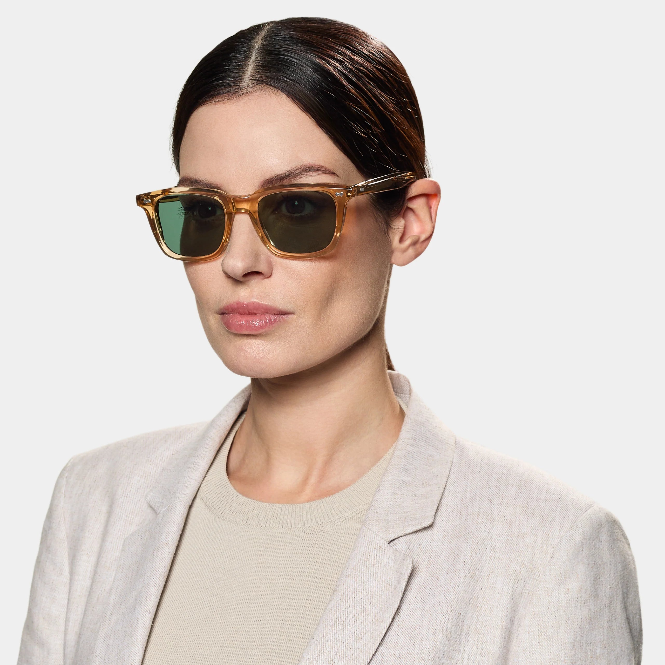 A woman with fair skin and dark, slicked-back hair wears TBD Eyewear's Carré Eco Champagne | Bottle Green sunglasses, paired with a light beige blazer and cream top, posing neutrally against a plain light gray background. , available at Richard Gelding Mayfair