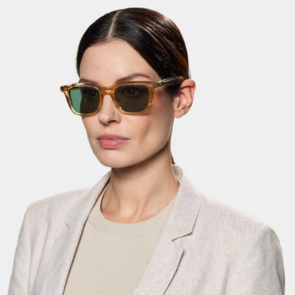 A woman with fair skin and dark, slicked-back hair wears TBD Eyewear's Carré Eco Champagne | Bottle Green sunglasses, paired with a light beige blazer and cream top, posing neutrally against a plain light gray background. , available at Richard Gelding Mayfair
