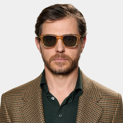 A man with short brown hair, a beard, and mustache wears TBD Eyewear's Carré Eco Champagne | Bottle Green sunglasses. He looks ahead with a neutral expression, dressed in a green collared shirt and brown houndstooth blazer against a white background. , available at Richard Gelding Mayfair