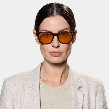 A woman with fair skin and dark hair slicked back wears TBD Eyewear’s Carré Eco Dark Havana | Orange oversized sunglasses and a light beige blazer over a cream top, facing the camera against a plain, light gray background. , available at Richard Gelding Mayfair