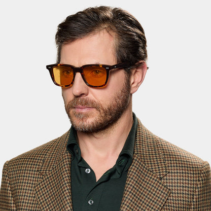 A man with short brown hair and a beard wears TBD Eyewear's Carré Eco Dark Havana | Orange sunglasses, a green collared shirt, and a brown plaid blazer, looking slightly to the side against a plain white background. , available at Richard Gelding Mayfair