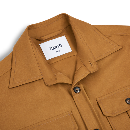 A close-up of the Manto Camel Wool/Silk Gubbio Overshirt shows a brown collared shirt with two front flap pockets and dark buttons. The thick, structured fabric blends premium wool and silk, reflecting quality materials and craftsmanship. , available at Richard Gelding Mayfair