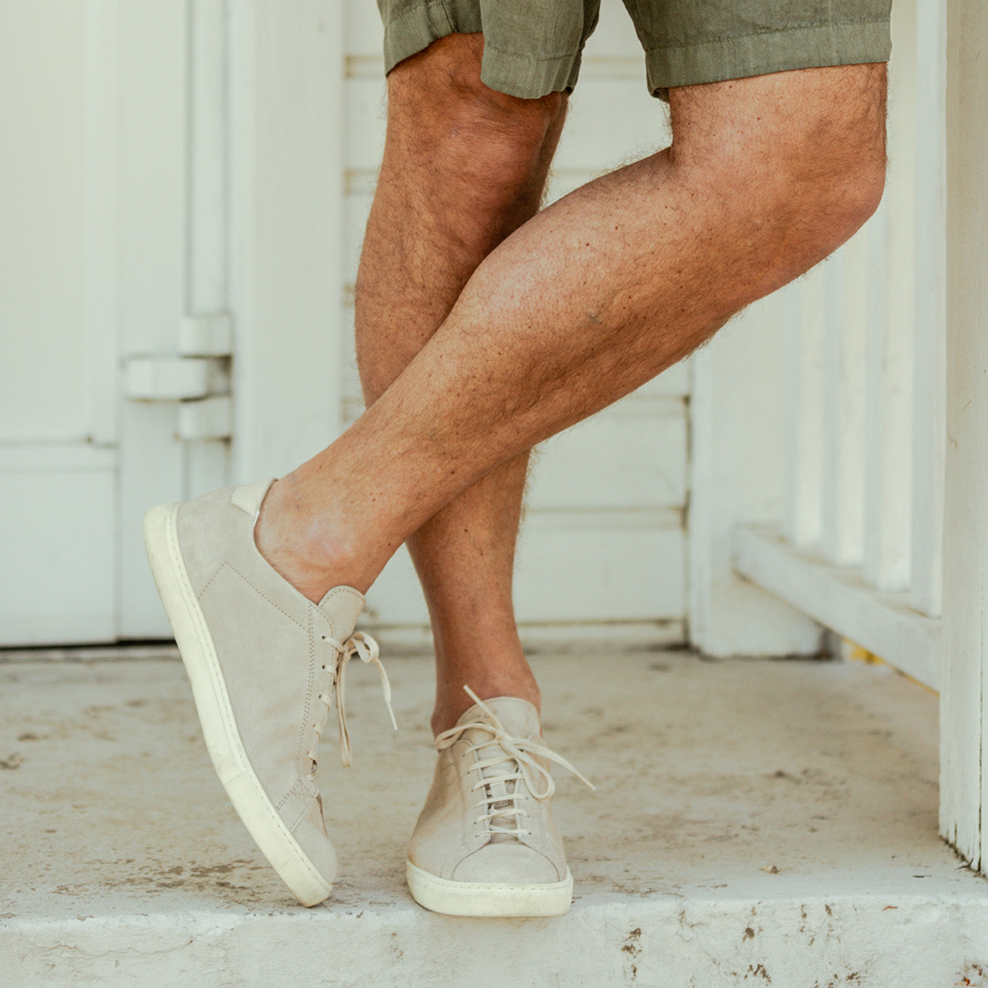 A man stands on a porch, crossing his ankles, wearing khaki shorts and Beige Suede Leather Racquet sneakers with Margom rubber soles by CQP.