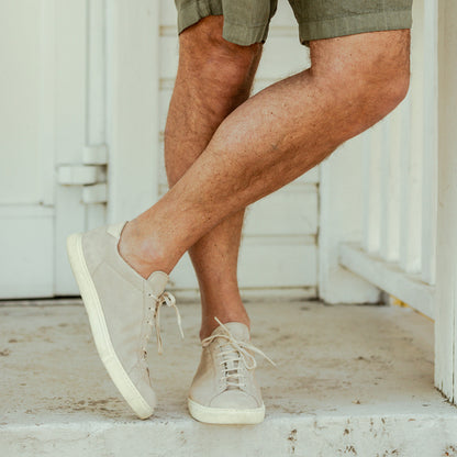 A man stands on a porch, crossing his ankles, wearing khaki shorts and Beige Suede Leather Racquet sneakers with Margom rubber soles by CQP.