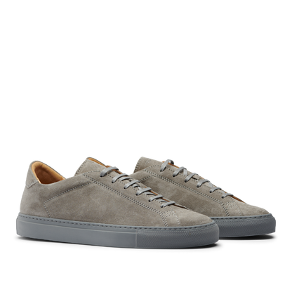 A pair of Granit Grey Suede Leather Racquet Sr Sneakers by CQP with white laces and a dark gray sole, displayed against a striped gray background.