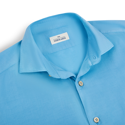 A neatly folded Blue Muslin Cotton Blend Beach Shirt by Alessandro Gherardi, featuring a pointed collar and beige buttons, is displayed on a white surface. , available at Richard Gelding Mayfair