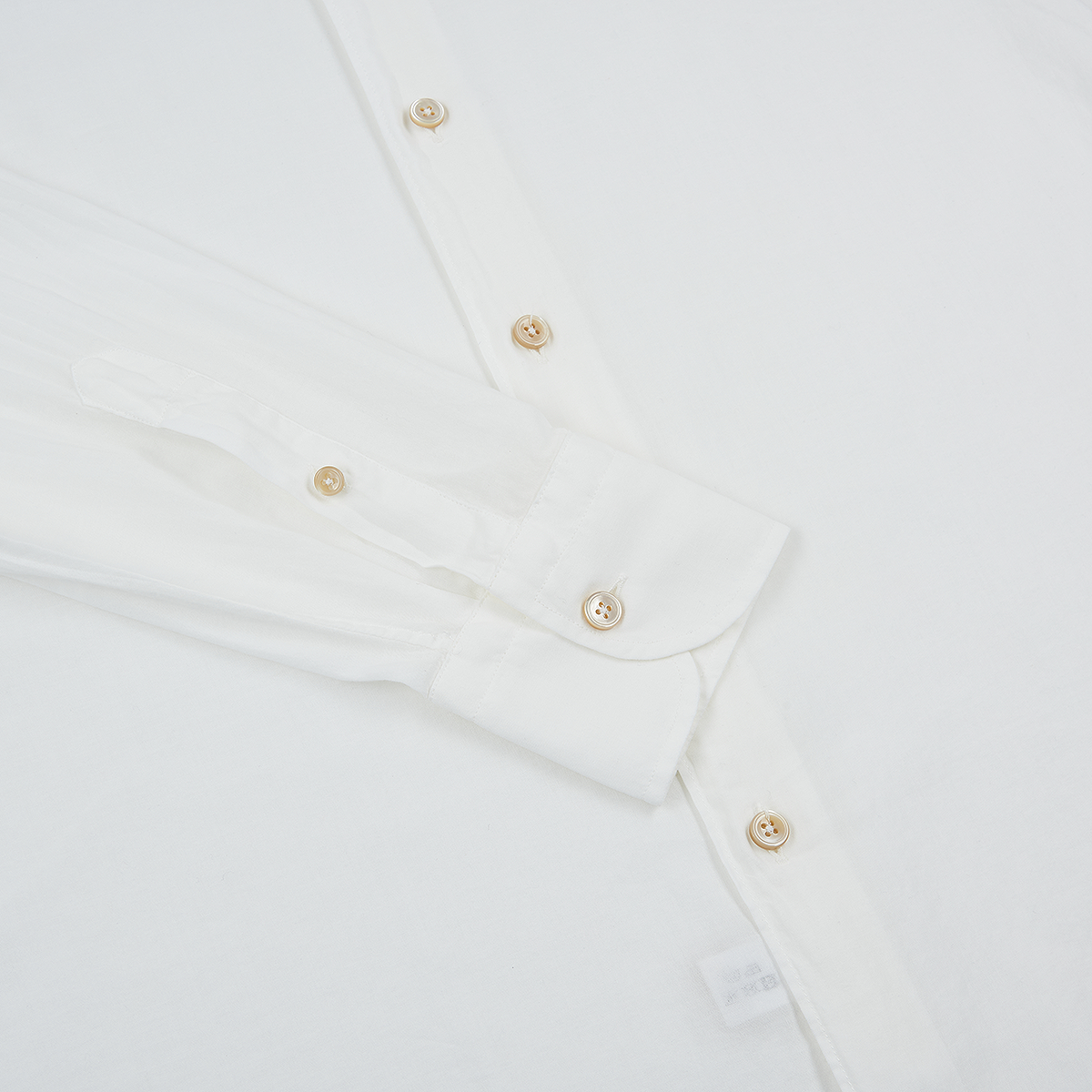 A close-up of the Alessandro Gherardi White Muslin Beach Shirt, neatly folded on a white surface. Cream-colored buttons, visible cuff and sleeve, part of the front placket, and a small white tag near the bottom edge are shown. , available at Richard Gelding Mayfair