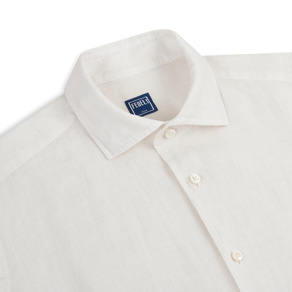 A close-up of the Fedeli Off White Linen Cutaway Collar Shirt shows its refined texture and classic cutaway collar with white buttons, featuring a blue “FEDELI” label inside, laid flat on a white background. , available at Richard Gelding Mayfair
