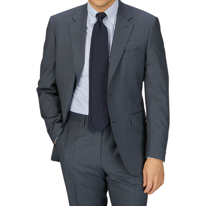 A man in a tailored Canali Grey-Blue Melange Wool Suit with a blue tie and striped shirt, posing with one hand in his pocket against a gray background.