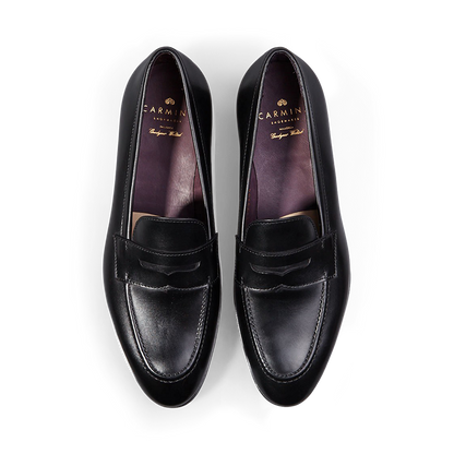 A pair of Carmina black calf leather Uetam penny loafers against a black background.