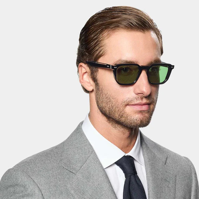 A man with short brown hair and stubble wears TBD Eyewear’s Dart Black | Green handcrafted Italian acetate sunglasses with green mineral glass lenses, a light gray suit jacket, white shirt, and navy tie against a plain light gray background. , available at Richard Gelding Mayfair