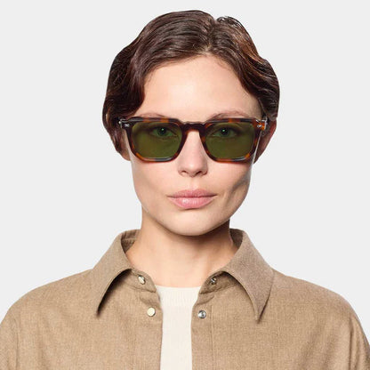 A person with short brown hair wears TBD Eyewear Dart Brown Havana | Green rectangular sunglasses, featuring green mineral glass lenses, and a beige collared shirt over a white top, against a plain light gray background, facing the camera with a neutral expression. , available at Richard Gelding Mayfair