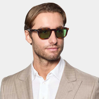 A man with light brown hair and a short beard wears TBD Eyewear Dart Brown Havana | Green sunglasses with recyclable acetate frames and green mineral lenses, styled with a beige pinstripe suit and white shirt, against a plain gray background. , available at Richard Gelding Mayfair