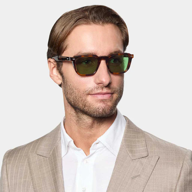 A man with light brown hair and a short beard wears TBD Eyewear Dart Brown Havana | Green sunglasses with recyclable acetate frames and green mineral lenses, styled with a beige pinstripe suit and white shirt, against a plain gray background. , available at Richard Gelding Mayfair