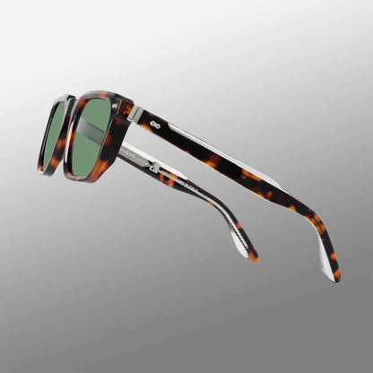The Dart Brown Havana | Green by TBD Eyewear features thick, tortoiseshell-patterned recyclable acetate frames, dark green mineral glass lenses, metal-accented temples, and a subtle logo near the hinge, displayed on a gradient gray background. , available at Richard Gelding Mayfair