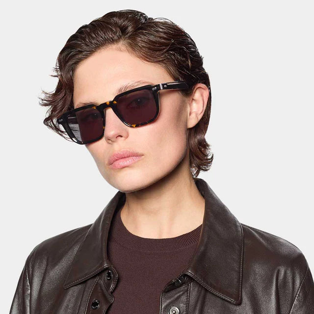 A person with short brown hair wears TBD Eyewear Dart Eco Black Havana | Grey sunglasses with acetate renew frames and 100% UV protection, plus a brown leather jacket over a dark shirt, against a plain light gray background, facing the camera neutrally. , available at Richard Gelding Mayfair