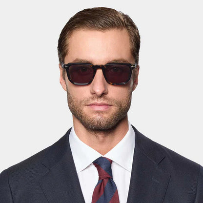 A man with short brown hair and a trimmed beard wears TBD Eyewear Dart Eco Black Havana | Grey sunglasses with 100% UV protection, a dark suit, white shirt, and red-navy striped tie against a plain light gray background. , available at Richard Gelding Mayfair