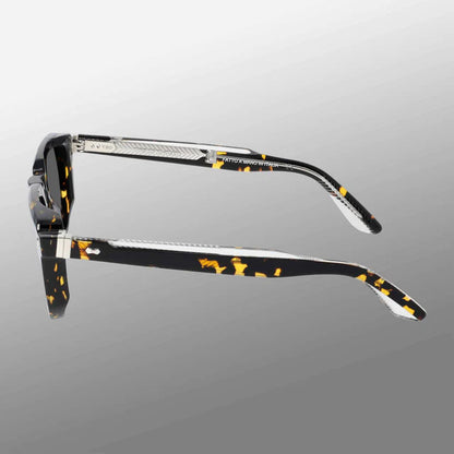 Side view of TBD Eyewear Dart Eco Black Havana | Grey sunglasses with acetate renew frames. The arms have metal detailing, textured accents, and a silver hinge, set against a smooth light-to-dark gray gradient. , available at Richard Gelding Mayfair