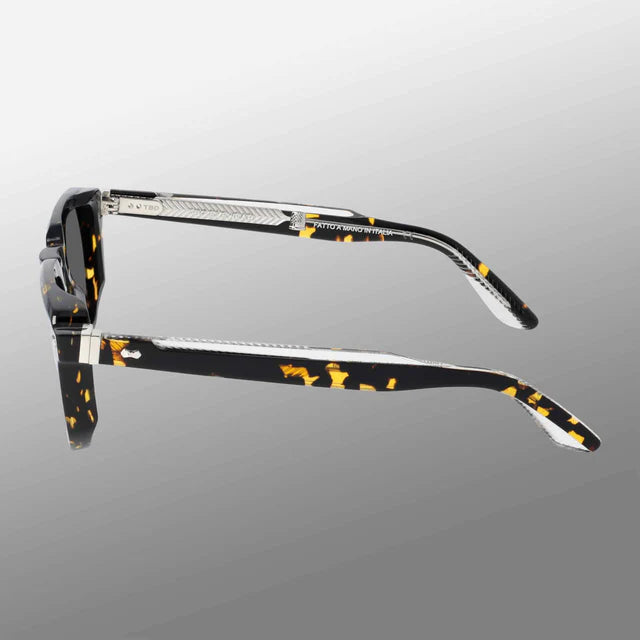 Side view of TBD Eyewear Dart Eco Black Havana | Grey sunglasses with acetate renew frames. The arms have metal detailing, textured accents, and a silver hinge, set against a smooth light-to-dark gray gradient. , available at Richard Gelding Mayfair
