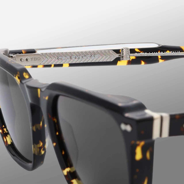 Close-up of TBD Eyewear Dart Eco Black Havana | Grey sunglasses with acetate renew frames, a black and yellow marbled design, silver hardware, engraved arm text, and 100% UV-protective dark lenses on a light gray gradient background. , available at Richard Gelding Mayfair