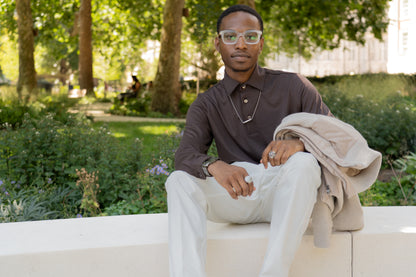 A man with short hair and glasses sits on a white bench in a green park, wearing the Herno Brown Wool Jersey Long Sleeve Polo, light pants, and jewelry. He holds a beige jacket and looks at the camera as sunlight filters through the trees, with a blurred figure seated in the background. , available at Richard Gelding Mayfair