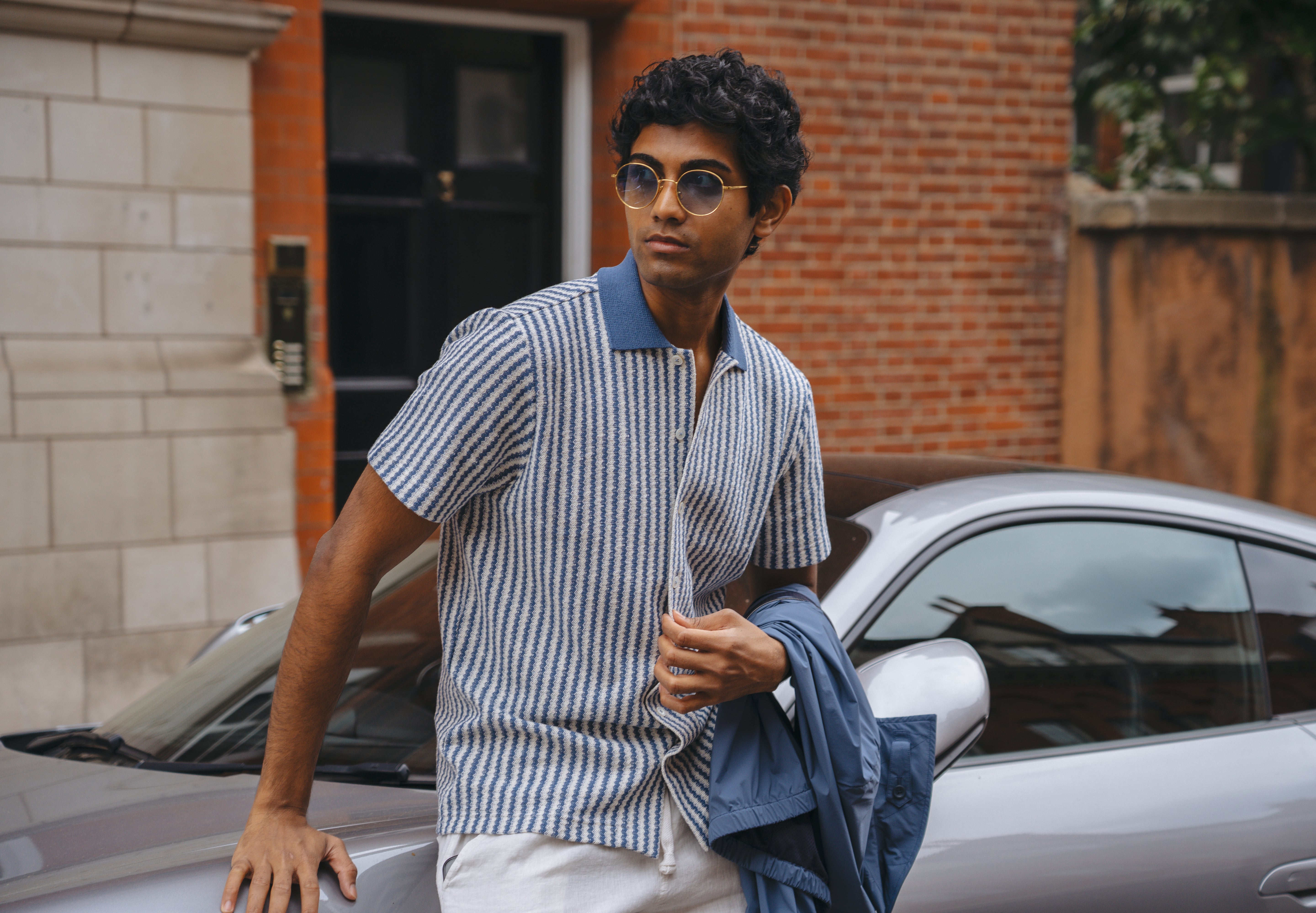 A man with curly hair wearing round sunglasses and the Gran Sasso Blue Stripe Button-Through Knit Polo stands by a silver car, holding a blue jacket. Brick and stone buildings with a black door are in the background as he looks to the side, relaxed. , available at Richard Gelding Mayfair