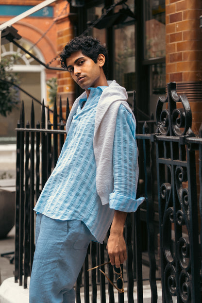 A young person with short curly hair leans on a black iron fence outdoors, wearing the Alessandro Gherardi Blue Stripe Cotton/Linen Popover Camp Collar Shirt, light blue pants, and a gray sweater over their shoulders, holding sunglasses and looking calmly at the camera. , available at Richard Gelding Mayfair