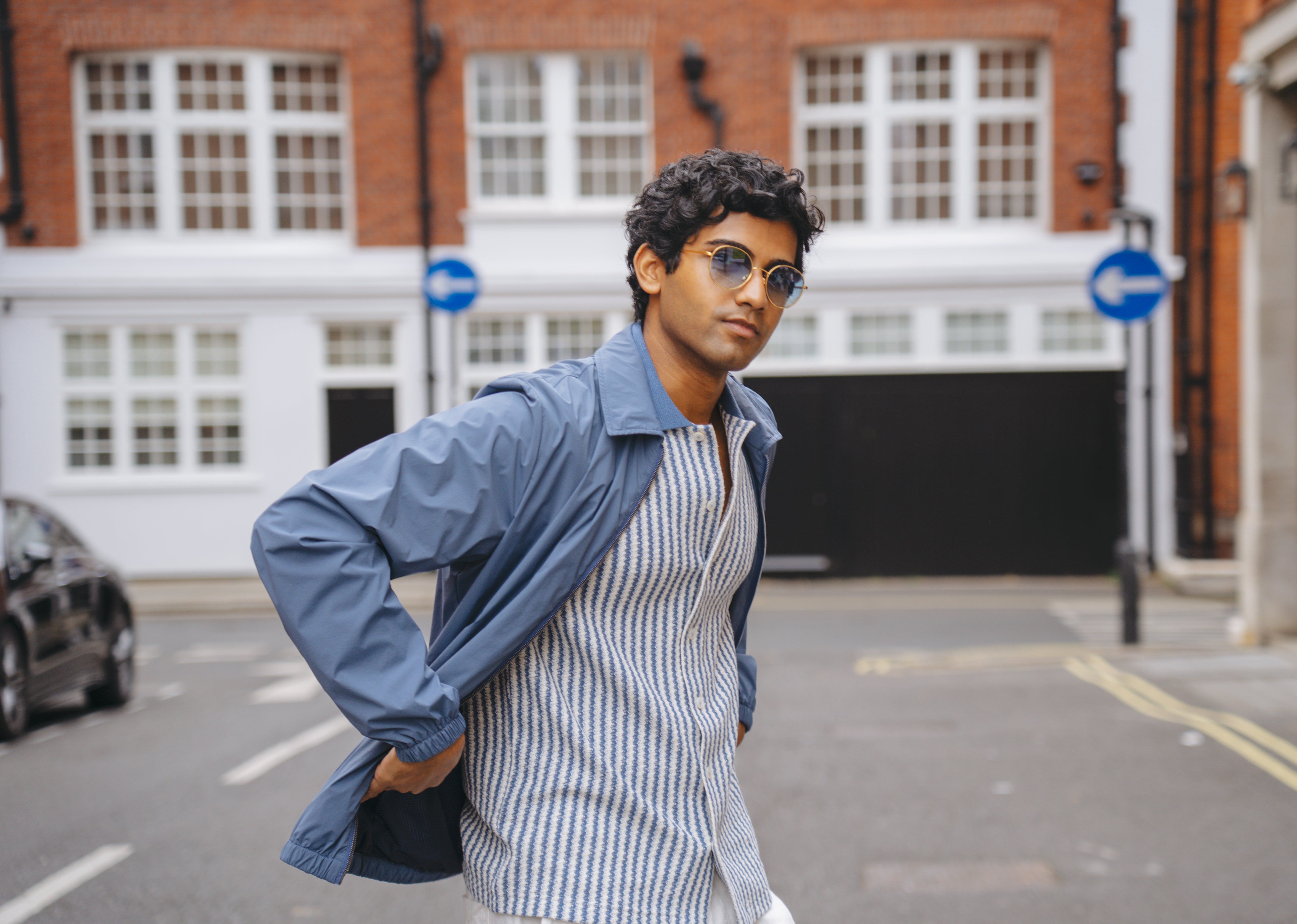 A man with dark, curly hair and sunglasses wears a Gran Sasso Blue Stripe Button-Through Knit Polo and a blue jacket as he walks down a city street lined with red-brick and white buildings, blue road signs, and an urban vibe. , available at Richard Gelding Mayfair