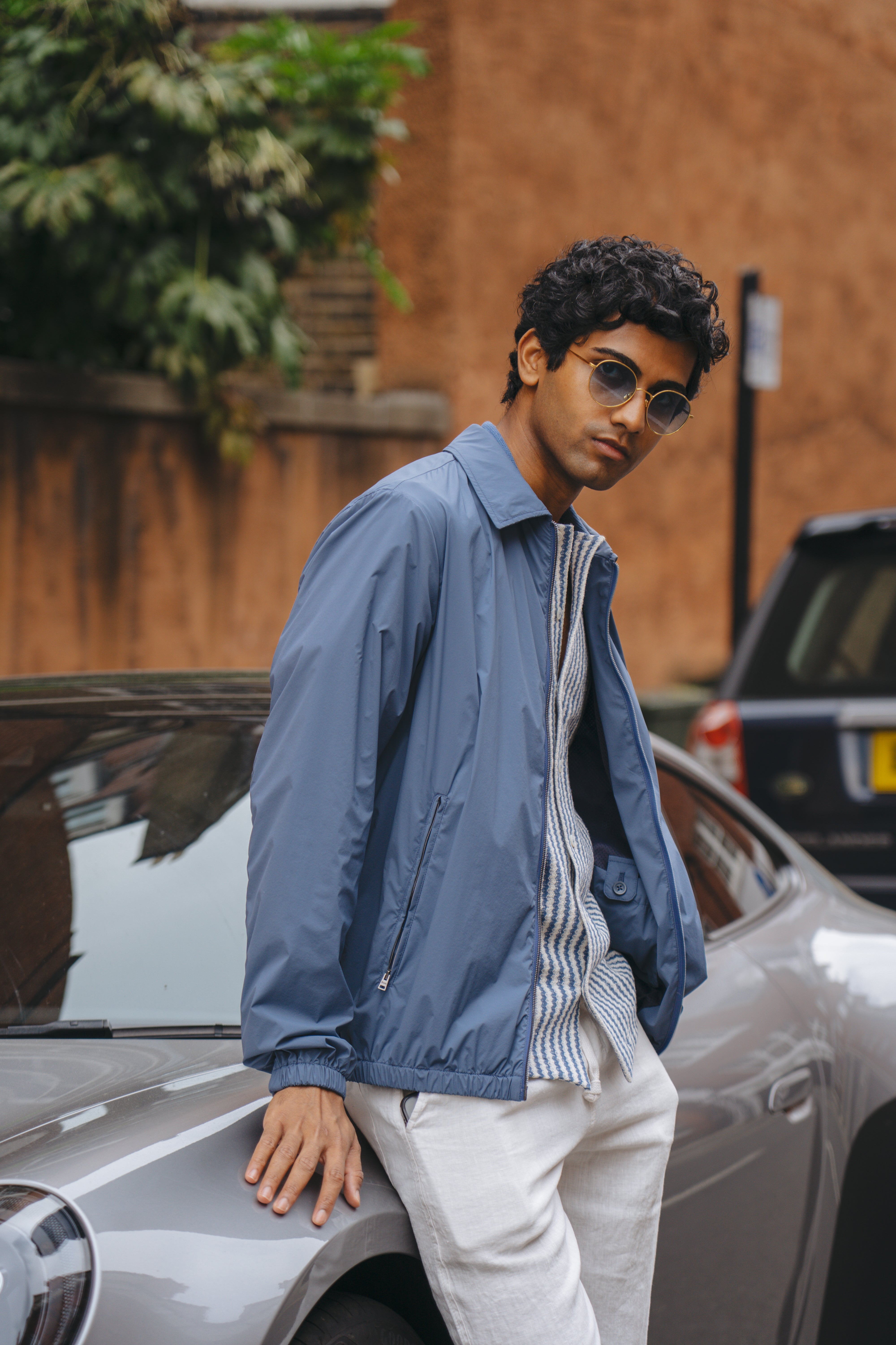 A young man in sunglasses and a Herno Blue Lightweight Nylon Zip Jacket leans against a sleek grey car, with a brown wall, wooden fence, foliage, and a partially visible black car in the background. , available at Richard Gelding Mayfair
