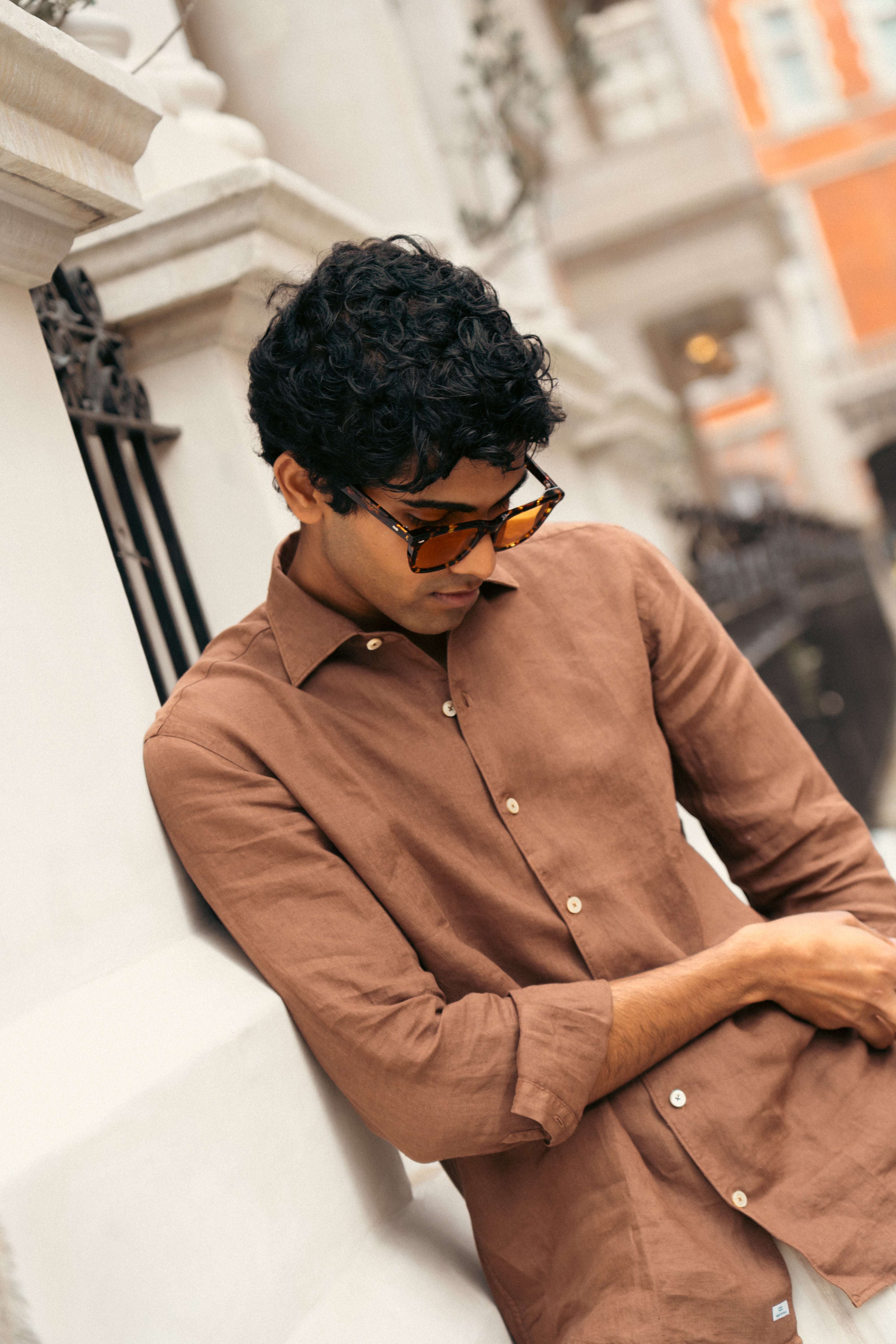A man with short, curly black hair wears tortoiseshell sunglasses and the Alessandro Gherardi Tobacco Brown Linen One Piece Collar Casual Shirt. He leans against a light building, looking down with a relaxed expression. Warm tones blur the background architecture. , available at Richard Gelding Mayfair