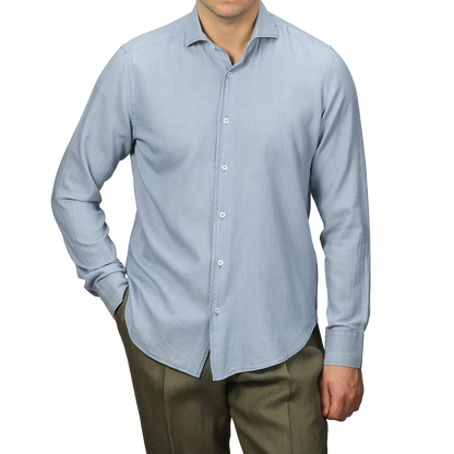 A person dressed in a Fedeli Light Blue Cotton Denim Shirt and olive green pants stands with one hand in their pocket against a plain gray background.