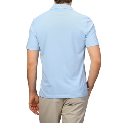 A person is seen from the back in a luxurious Fedeli light blue Capri collar polo shirt, crafted from organic cotton, paired with beige pants against a plain backdrop.