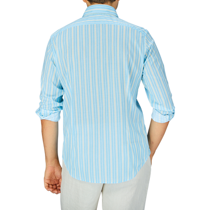 A person standing with their back to the camera, wearing a Fedeli Sky Blue Yellow Striped Cotton Beach Shirt and white pants.