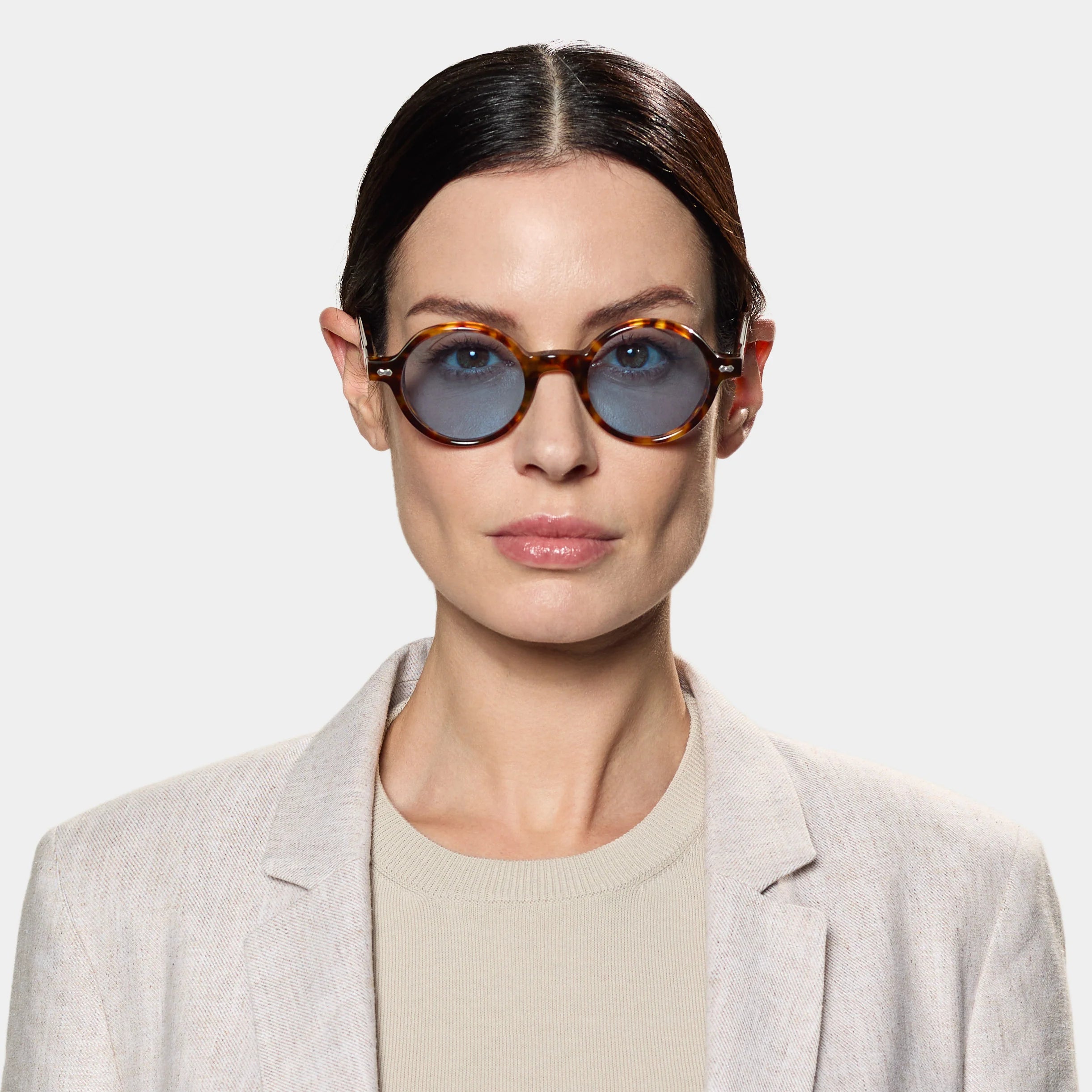 A woman with dark hair tied back wears TBD Eyewear's Giotto Eco Spotted Havana | Blue sunglasses—round, brown tortoiseshell frames with blue lenses—paired with a beige blazer and crewneck top against a white background. , available at Richard Gelding Mayfair