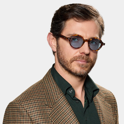 A man with short brown hair and a beard wears TBD Eyewear's Giotto Eco Spotted Havana | Blue round sunglasses, a brown and tan houndstooth blazer, and a dark green shirt. He poses neutrally against a plain white background. , available at Richard Gelding Mayfair