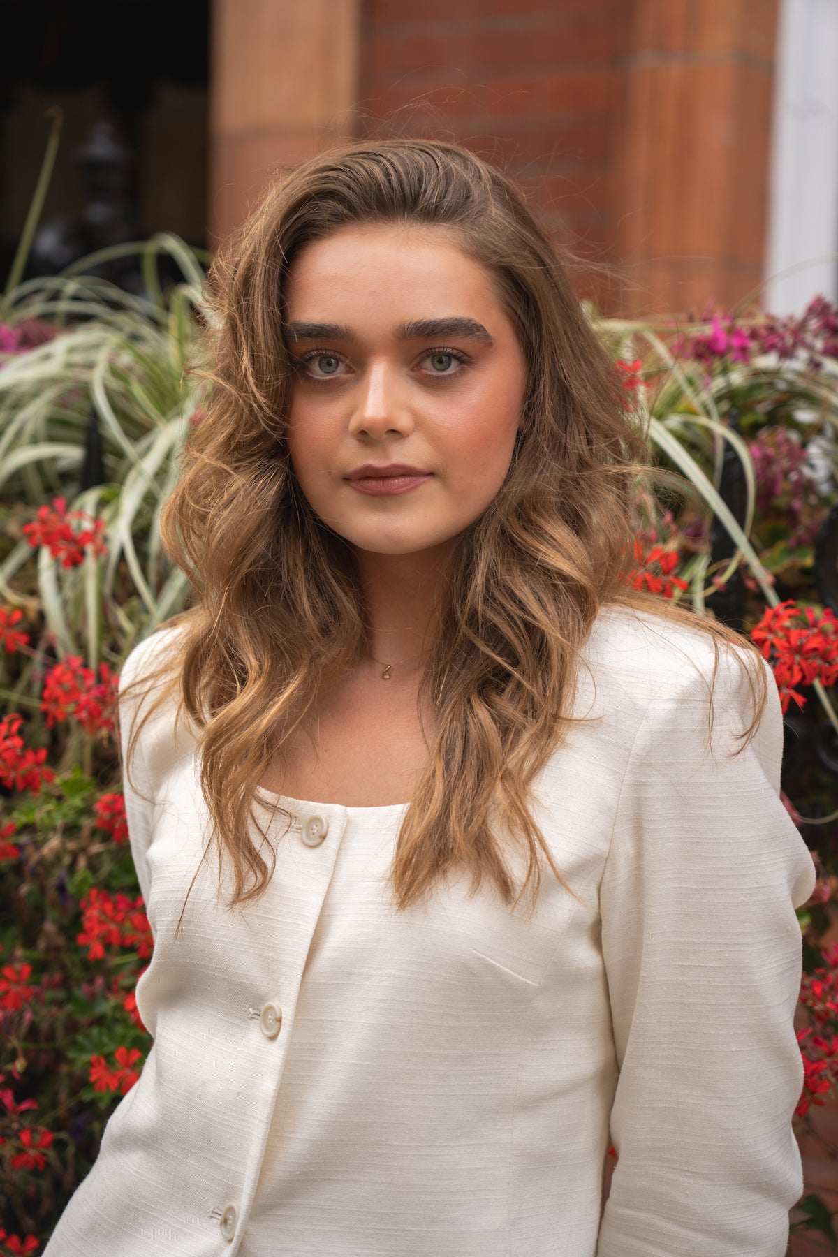 A woman with wavy light brown hair wearing a cream-colored textured jacket, standing outdoors in front of a brick wall and green plants with red flowers.