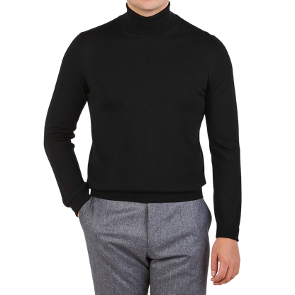 A person wears the Gran Sasso Black Extra Fine Merino Wool Rollneck with gray trousers, standing with one hand in their pocket against a plain background.
