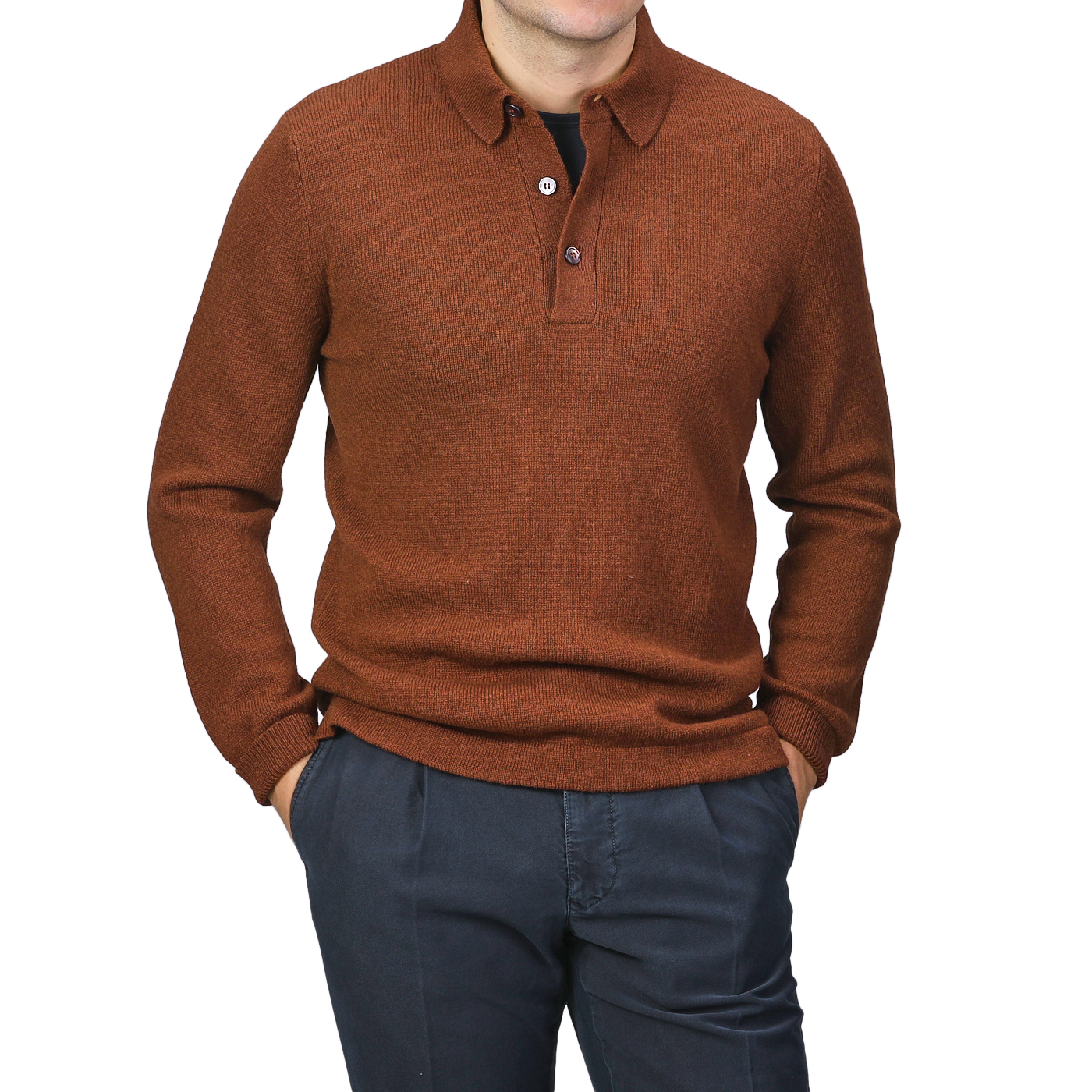 A man models the Burnt Orange Virgin Wool LS Polo Shirt from Gran Sasso, styled with dark pants and hands in his pockets against a neutral background.