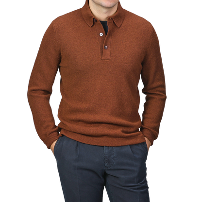 A man models the Burnt Orange Virgin Wool LS Polo Shirt from Gran Sasso, styled with dark pants and hands in his pockets against a neutral background.